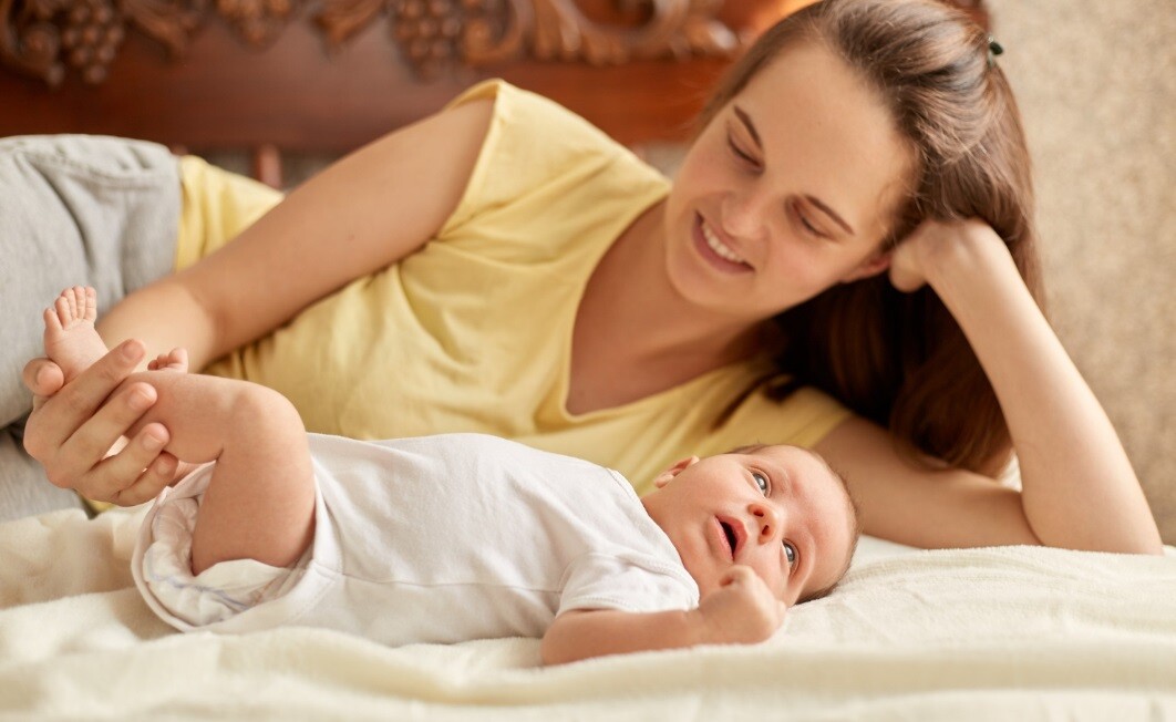 a mother in yellow shirt lying near to her newborn baby dressed in white. They both are on the bed.