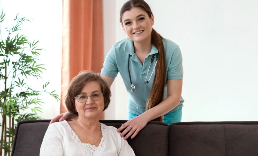 an elderly lady sitting on a sofa and a young lady standing adjacent to her. Behind them is a white wall.