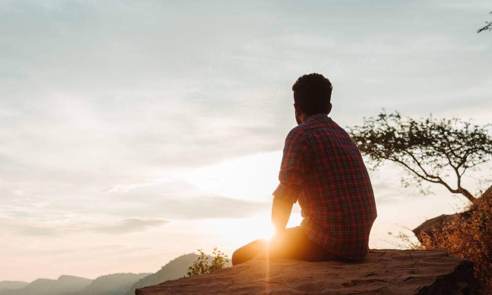 A man in a plaid shirt peacefully sits on a rock and gazes into the distance as the sun sets in the sky.