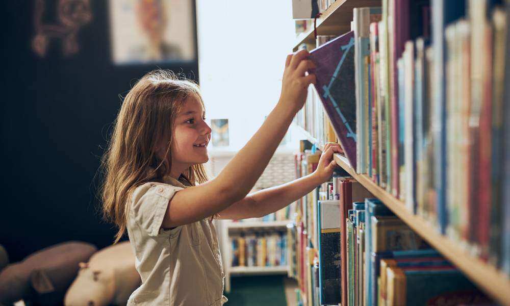 An elementary-aged girl smiles as she pulls a purple picture book from a well-stocked bookshelf in a library.