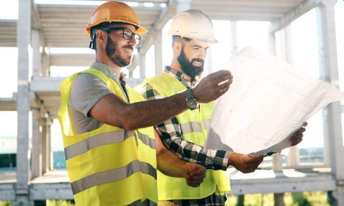 Two men in hard hats and high-visibility vests smile as they look at a large sheet of paper on a construction site.