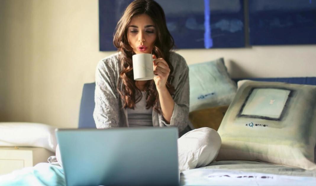 a lady drinking a cup of tea in front of the laptop and on top of the bed at home