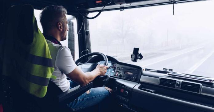 A truck driver behind the wheel of his semi, wearing a comfortable white T-shirt and jeans. He's focused on the road.