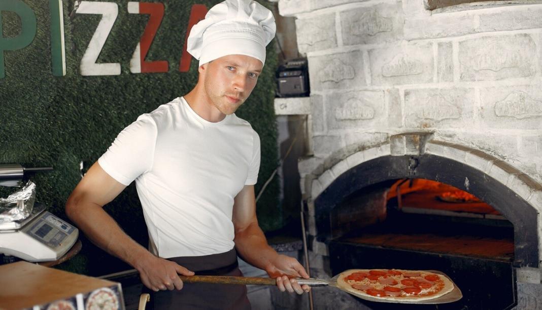 a man holding a pizza on a pane taking it to a burning furnace
