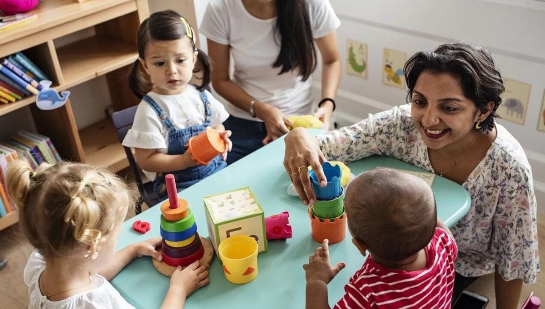 a kid's teacher is sitting around a table with four children as if giving speech therapy