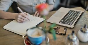 a lady writing on a diary with a laptop placed on the table