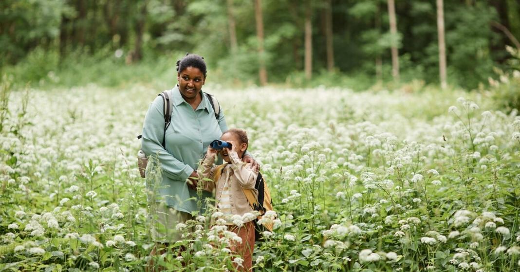 A Black mother with her young daughter in a field of flowers birdwatching. The daughter has a pair of binoculars.
