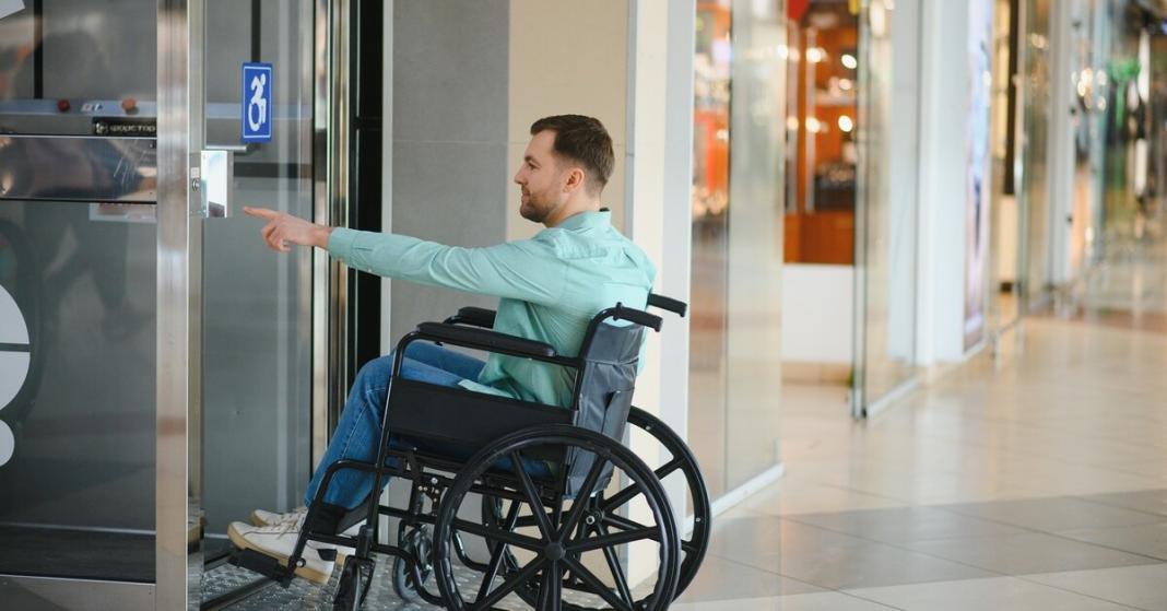 How To Navigate Public Spaces With a Wheelchair A man wearing a mint green shirt and jeans sits in a wheelchair. He's about to press a button on a door.