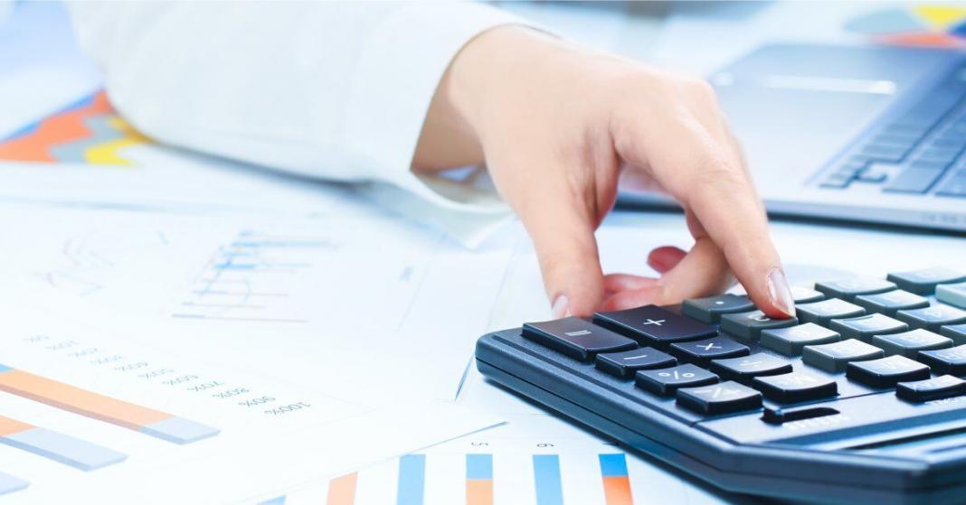 A close-up on a person's hands holding a pen and tapping on a calculator with various charts and graphs spread out.