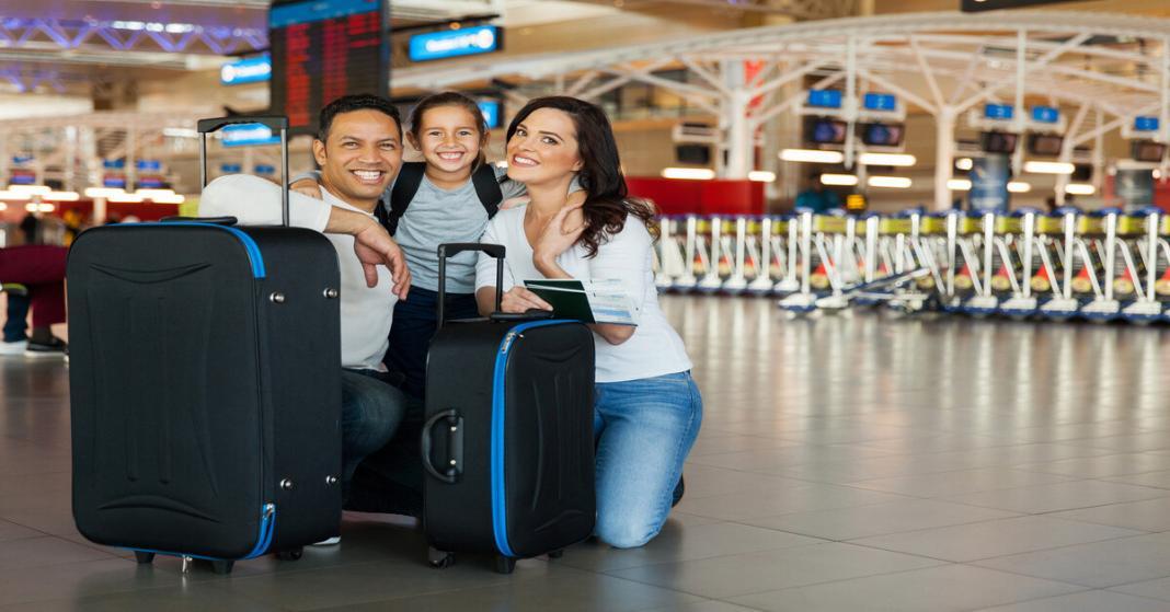 A family of three kneels down next to their luggage and smiles in the middle of an airport. A row of carts sits behind them.