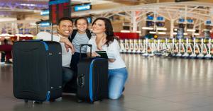 A family of three kneels down next to their luggage and smiles in the middle of an airport. A row of carts sits behind them.