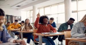 A college student is raising her hand in a classroom filled with other students. They are sitting at wooden desks.