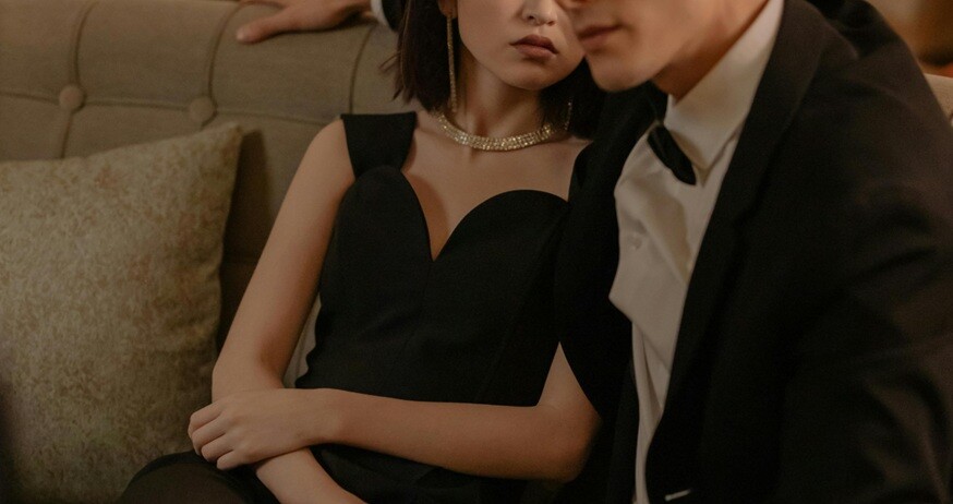 a lady in black dress wearing a diamond necklace and an handsome man sitting beside