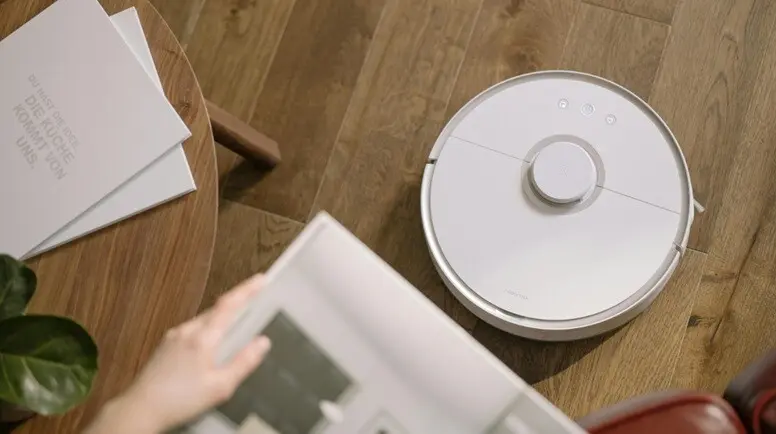 a robot cleaner seen a wooden floor with a lady reading newspaper in a sofa