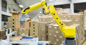A yellow robotic arm holding up a folded box in front of a dispenser with pallets of stacked cardboard boxes behind it.