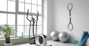 A home gym contains an elliptical, two silver yoga balls, two tennis rackets hanging on the wall, teal sneakers, and yoga mats.