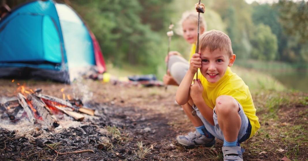 A boy wearing a yellow t-shirt, squatting while holding a stick with a marshmallow next to a camping tent.