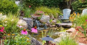 A garden with pink, orange, white, and yellow flowers, around a small pond with a small waterfall and two chairs.