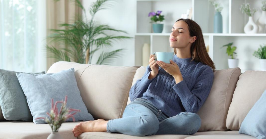 A woman sitting on a beige couch with blue pillows, holding a cup with both hands, inside a room with white walls.