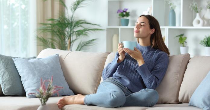 A woman sitting on a beige couch with blue pillows, holding a cup with both hands, inside a room with white walls.