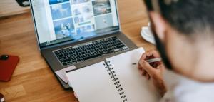 person writing on a notepad by looking a laptop with images on it