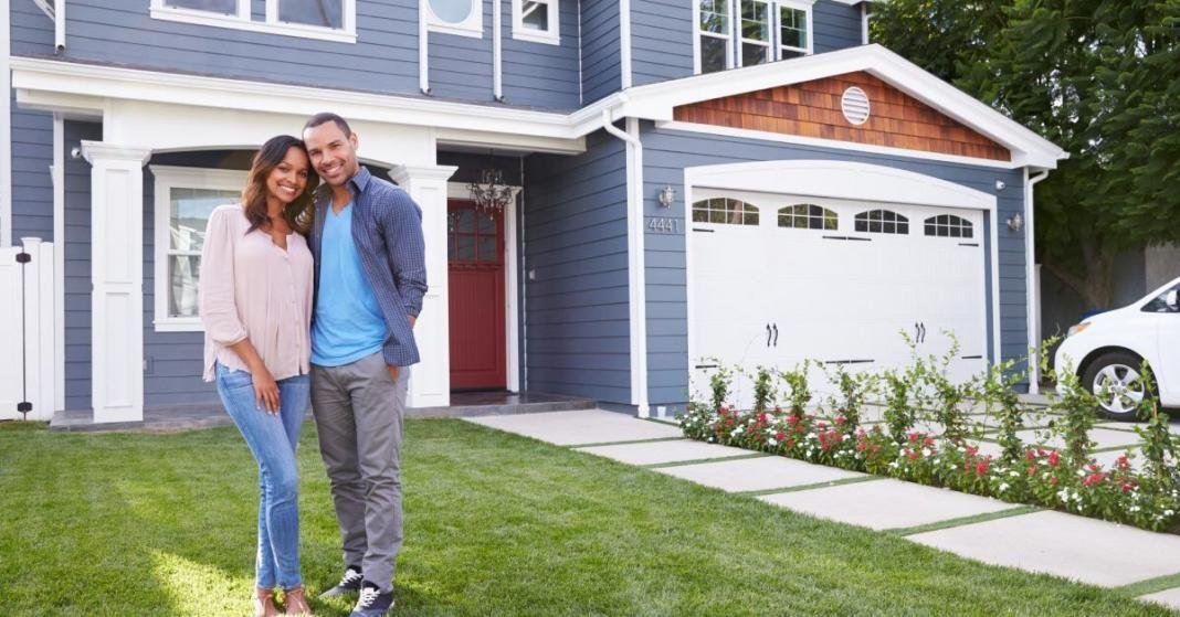 A man and a woman pose on the lawn in front of a two-story home with blue siding, white trim, and a brown front door.