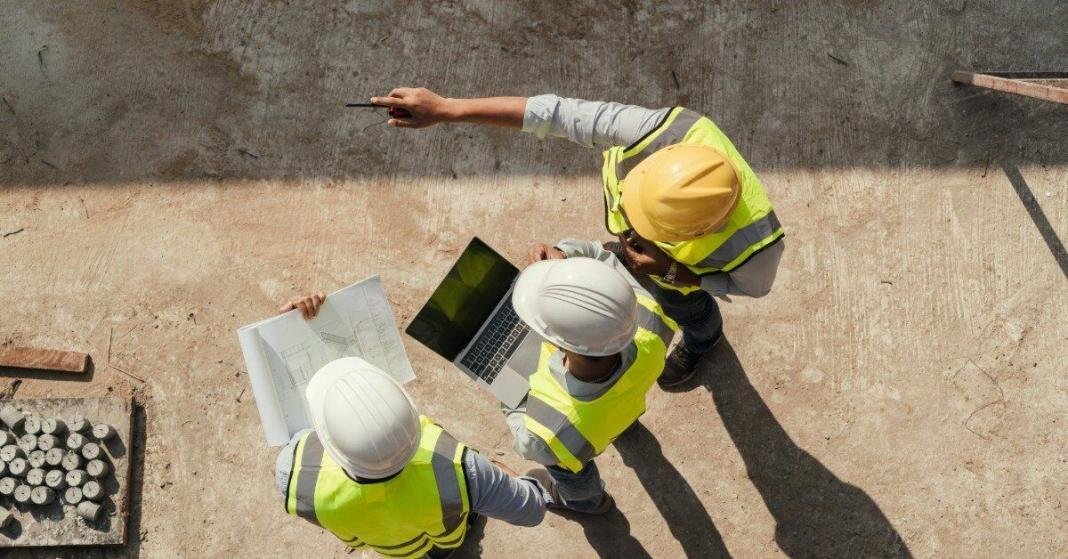 Three industrial site engineers stand together to discuss project plans. One points at the job and another holds a laptop.