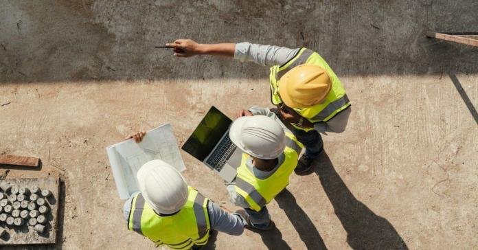 Three industrial site engineers stand together to discuss project plans. One points at the job and another holds a laptop.