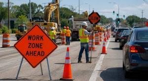 Road construction work zone with traffic control barriers, safety cones, construction workers in reflective gear, and vehicles moving through an organized roadway project under daylight conditions.