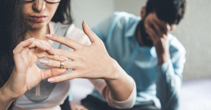 A close-up of the hand of a woman taking off her wedding ring with a man sitting behind her holding his face.