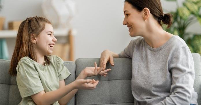 A mother and a young girl smile and talk on a gray couch in a bright living room with plants nearby.