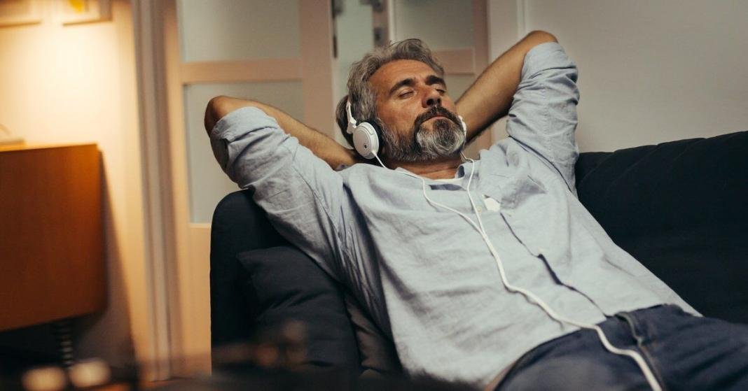 A middle-aged person relaxing on a lounging chair at home while listening to music using over-ear headphones.