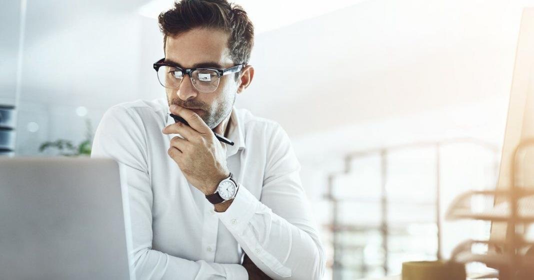 A man in glasses looks at a laptop while holding a pen at a bright office desk with an open notebook nearby.