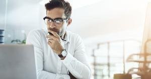 A man in glasses looks at a laptop while holding a pen at a bright office desk with an open notebook nearby.