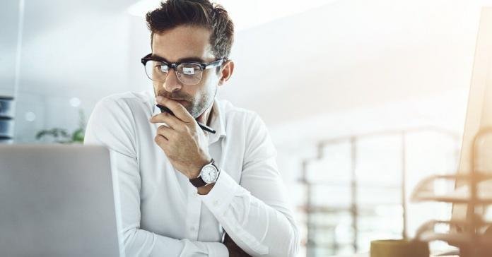 A man in glasses looks at a laptop while holding a pen at a bright office desk with an open notebook nearby.