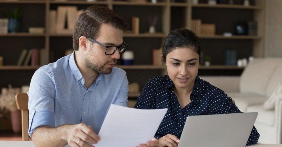 A man and a woman review a printed document beside a laptop while working together at a table in an office.