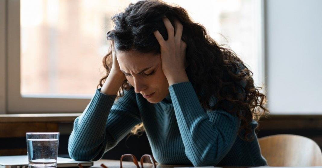 A woman is sitting at a desk as she holds her head in her hands. She looks distraught and in pain.