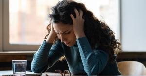 A woman is sitting at a desk as she holds her head in her hands. She looks distraught and in pain.