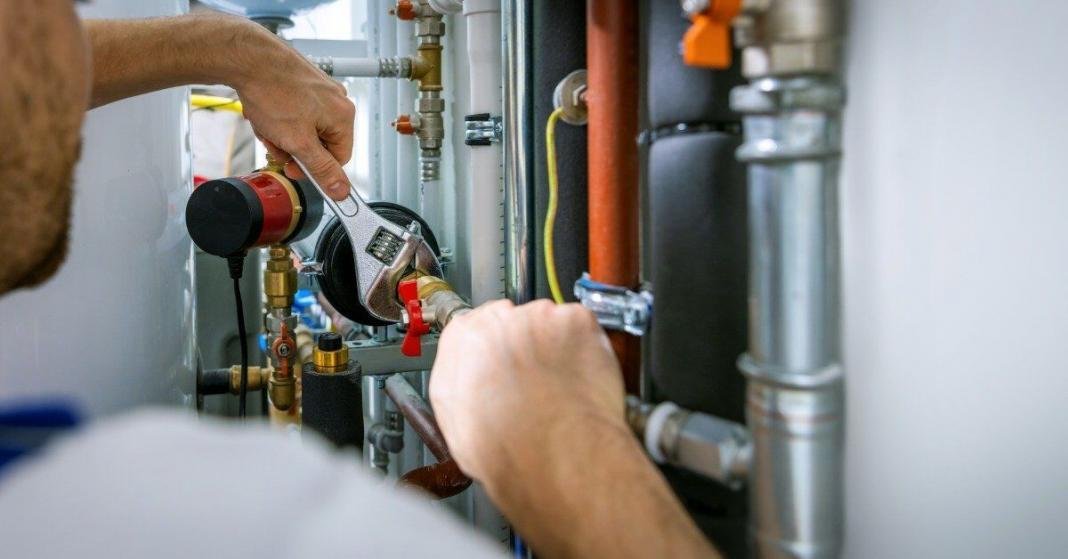 A plumber using an adjustable wrench on pipe fittings beside metal pipes and plumbing components in a utility space.