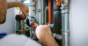 A plumber using an adjustable wrench on pipe fittings beside metal pipes and plumbing components in a utility space.
